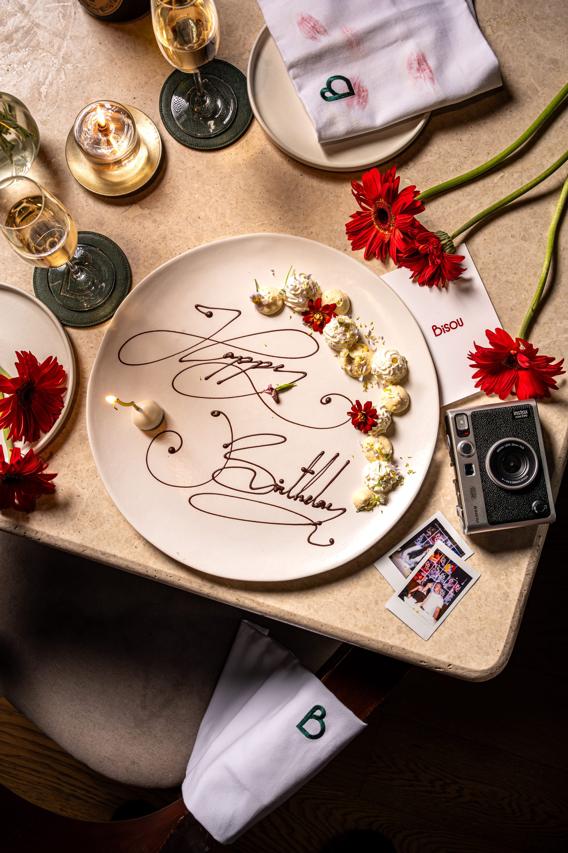 The birthday dessert plate at Bisou Bangkok: Happy Birthday piped in dark chocolate, quenelles, edible flowers, a lit candle, beside an instant camera and photographs