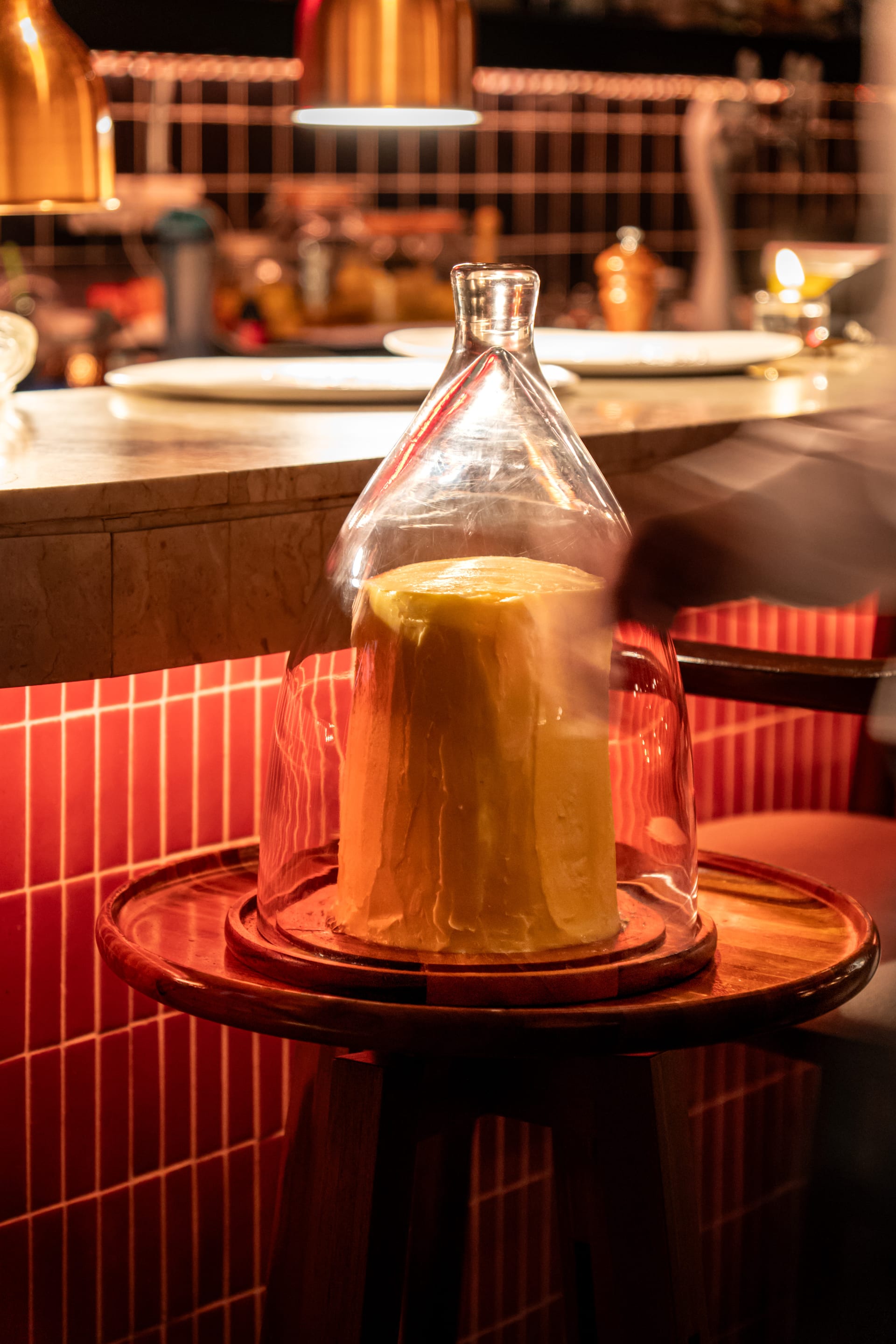 A whole block of French butter under a glass cloche on a wooden stand at Bisou Bangkok, the red-tiled bar behind it