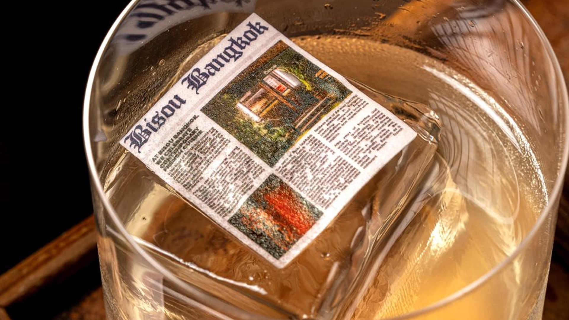 The New Year's Eve cover for Bisou Bangkok, a modern French restaurant on Soi Langsuan: an amber cocktail over a large clear ice cube printed with the Bisou Bangkok wordmark, on a wooden tray in the dark bar